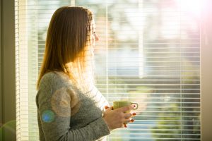 Woman standing at home by the window with cup of hot coffee wearing knitted warm sweater. Cozy sunny room filled with light. beautiful morning behind the big window.