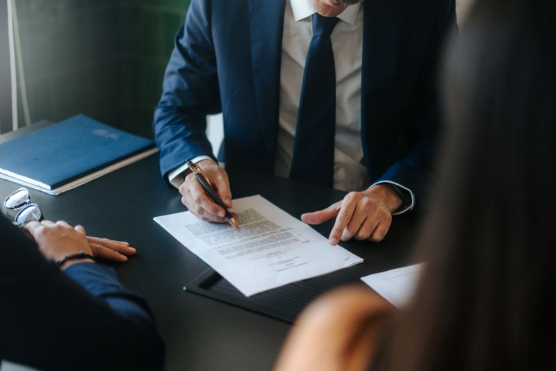 Two people sit across a desk from a lawyer who is writing on a legal document.